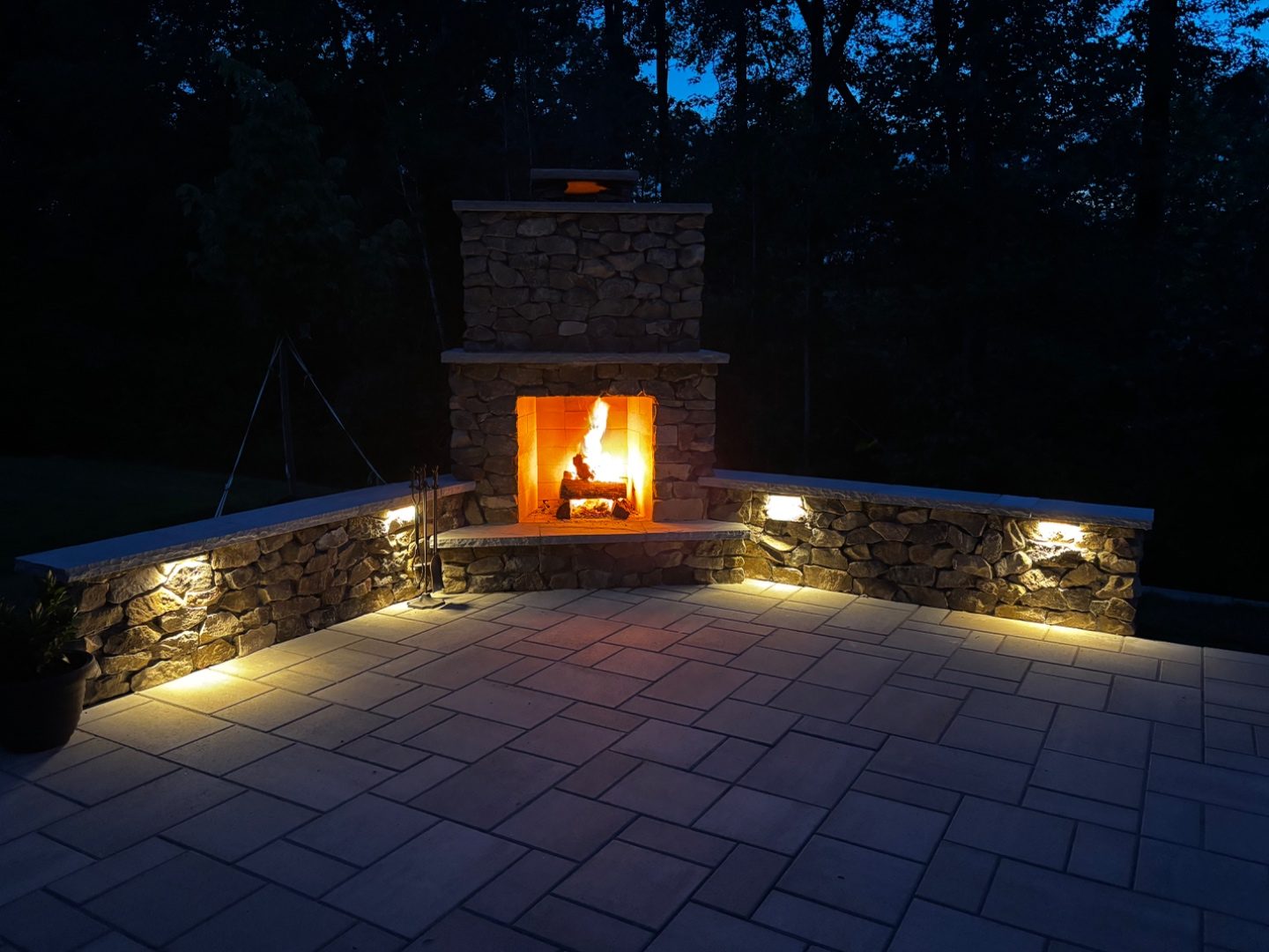 Illuminated stone outdoor fireplace and paver patio at dusk by Andrew's Landscaping